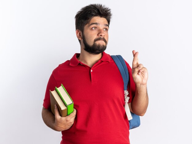 Young bearded student guy in red polo shirt with backpack holding notebooks making wish with hope expression crossing fingers standing