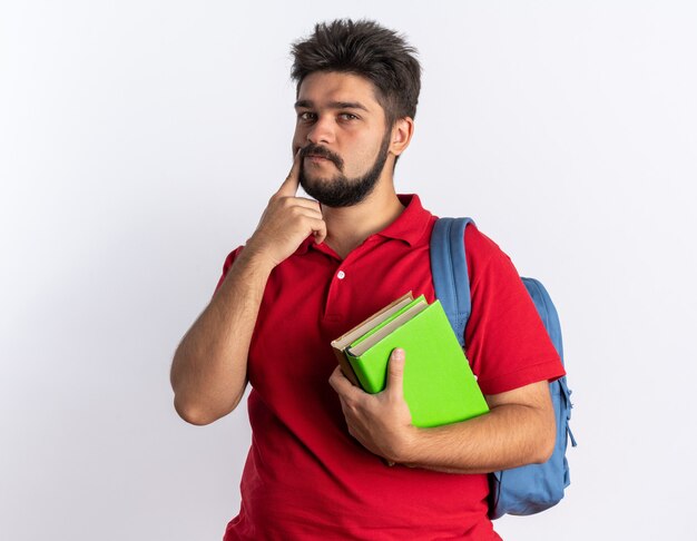 Young bearded student guy in red polo shirt with backpack holding notebooks  puzzled standing over white wall