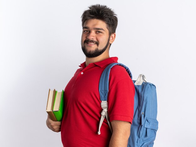 Young bearded student guy in red polo shirt with backpack holding notebooks  smiling confident happy and positive standing over white wall