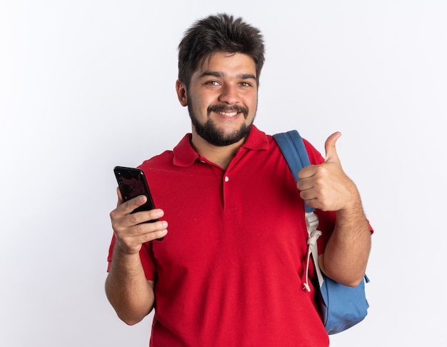 Young bearded student guy in red polo shirt with backpack holding smartphone  smiling cheerfully showing thumbs up standing over white wall