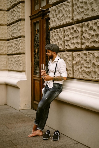 Free Photo young bearded tattooed man. a romantic guy in a white shirt, cap and suspenders walks in the city. revolves around a lamppost. peaky blinders. old-fashioned, retro.