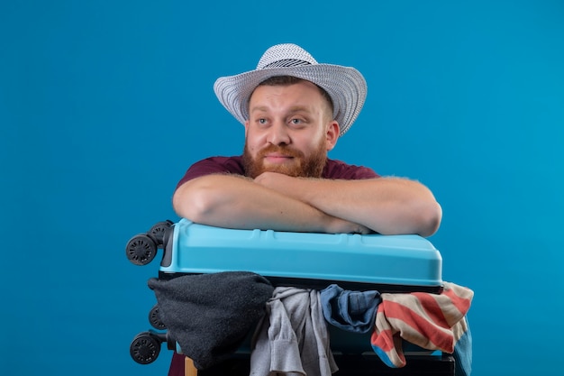 Free Photo young bearded traveler man in summer hat  with suitcase full of clothes optimistic and happy smiling looking aside with dreamy look 