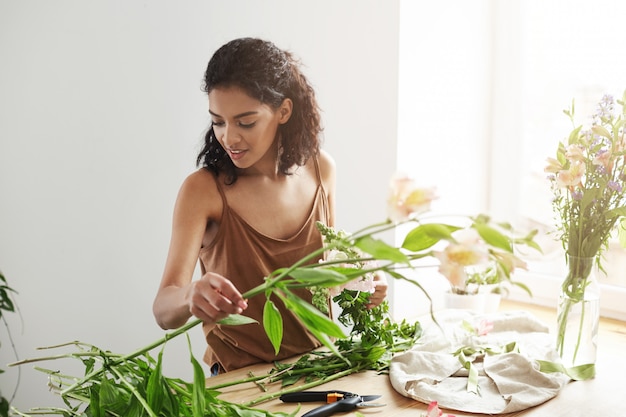 Free photo young beautiful african woman florist taking care of flowers at workplace over white wall.