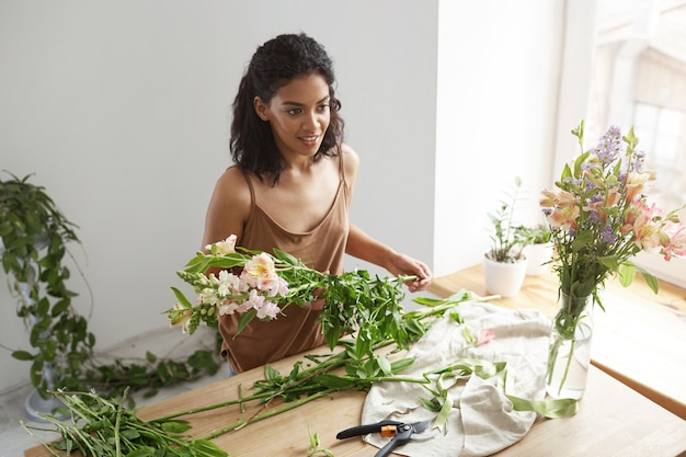 Young beautiful african woman florist taking care of flowers at workplace over white wall.