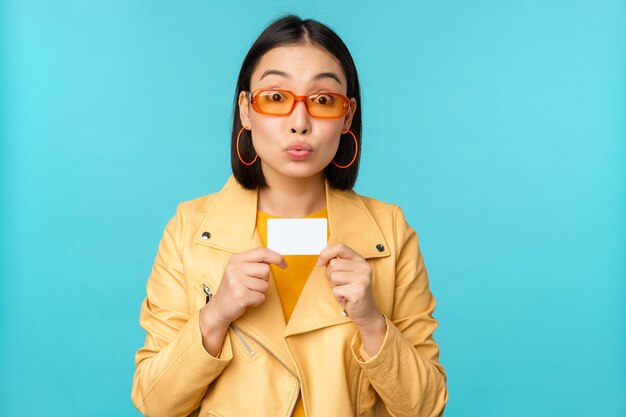 Young beautiful asian woman showing credit card smiling choosing bank standing over blue background Copy space