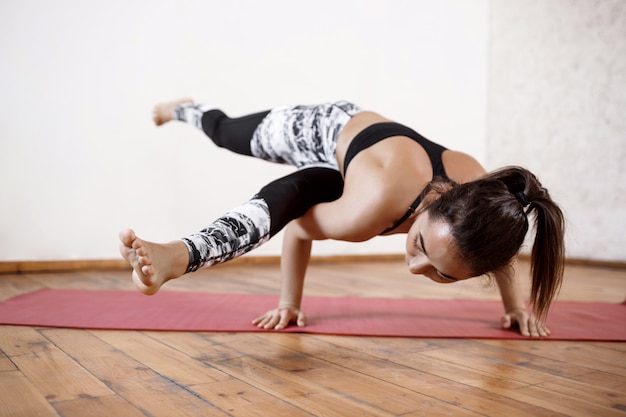 Young beautiful athletic woman practicing indoor yoga arm-balance Eka Pada Koundiyanasana