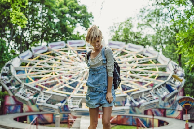 young beautiful blond girl in denim overall with a backpack posing in an amusement park