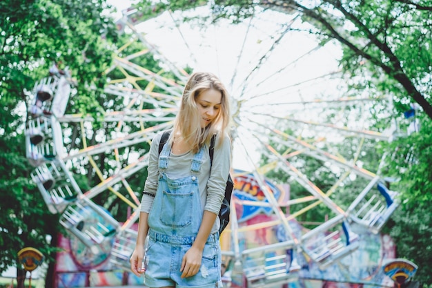 Free photo young beautiful blond girl in denim overall with a backpack posing in an amusement park