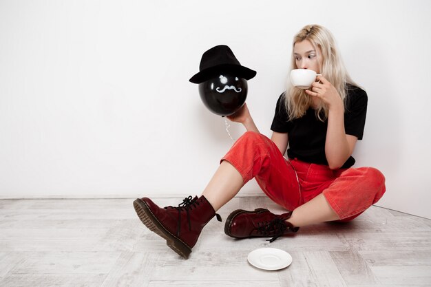 Young beautiful blonde girl holding black baloon in hat sitting on floor drinking coffee over white wall.