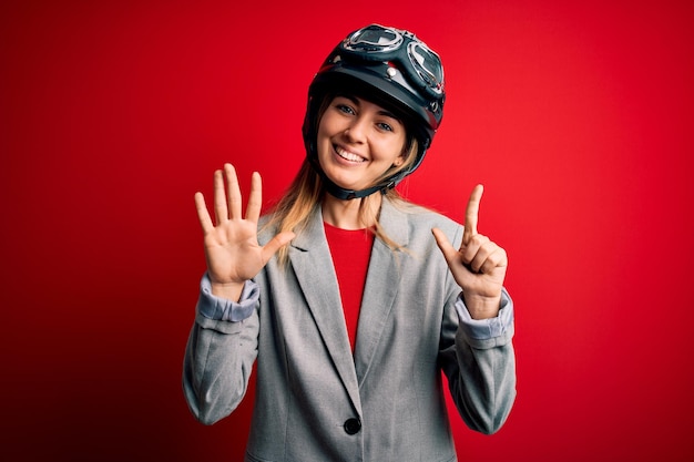 Free photo young beautiful blonde motorcyclist woman wearing motorcycle helmet over red background showing and pointing up with fingers number seven while smiling confident and happy