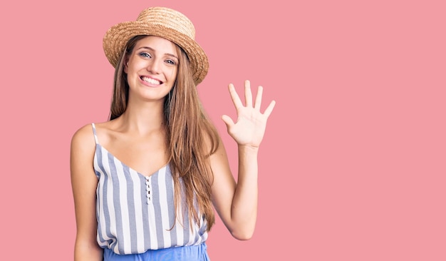 Young beautiful blonde woman wearing summer hat showing and pointing up with fingers number five while smiling confident and happy.