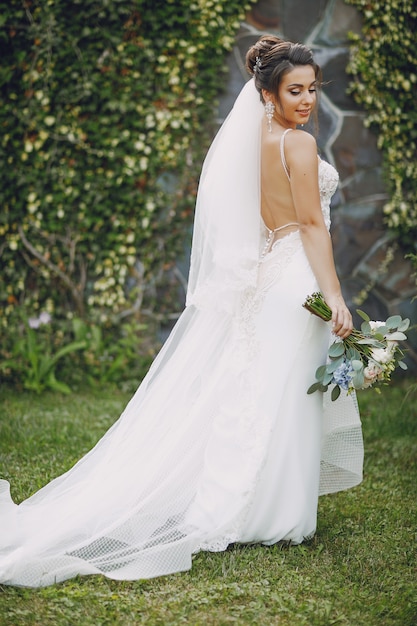 A young and beautiful bride is standing in a summer park with bouquet of flowers