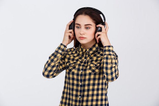 Young beautiful brunette girl dressed in plaid shirt listening music in headphones looking away over white wall