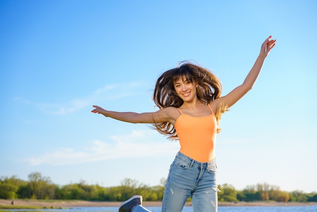 A young beautiful brunette girl in jeans and an orange t shirt jumps against the sky on a sunny summer day