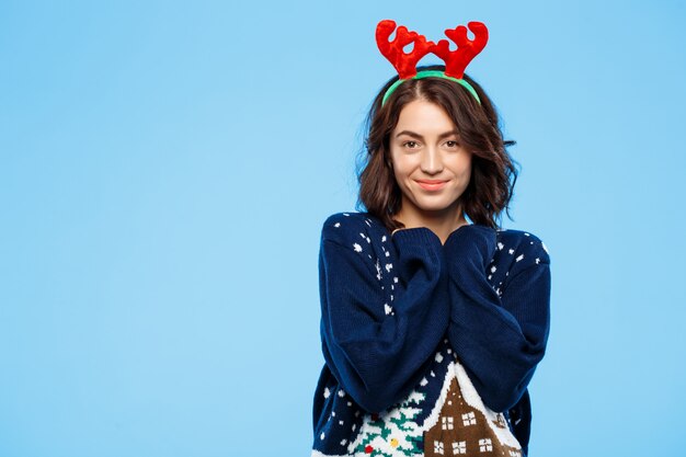 Young beautiful brunette girl in knitted sweater and christmas reindeer antlers smiling  over blue wall