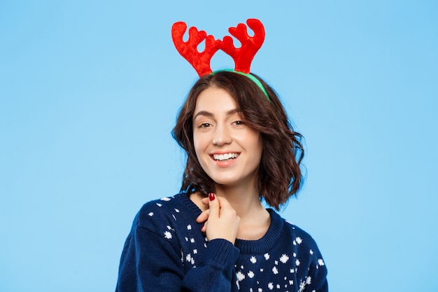 Young beautiful brunette girl in knitted sweater and christmas reindeer antlers smiling  over blue wall