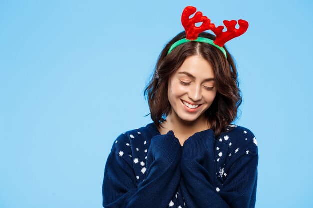 Young beautiful brunette girl in knitted sweater and christmas reindeer antlers smiling  over blue wall