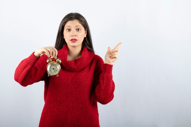 Young beautiful brunette woman holding an alarm clock and pointing away 