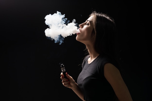 Free photo young beautiful brunette woman smoking, vaping e-cigarette with smoke on the black wall