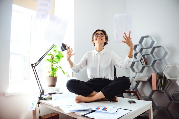 Young beautiful businesswoman in glasses smiling, throwing up papers, sitting on table at workplace in office.