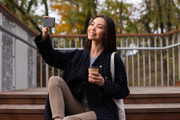 Young beautiful casual woman with coffee to go happily taking selfie on cellphone in autumn park