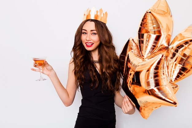 Young beautiful celebrating woman in black dress smile and posing with cocktail in hand and purity balloons .