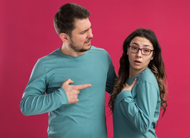 Young beautiful couple in blue casual clothes confused man pointing with index figner at his surprised girlfriend standing over pink wall