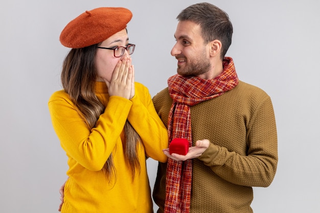 Free photo young beautiful couple happy man making proposal with engagement ring in red box to his amazed girlfriend in beret during valentines day standing over white background