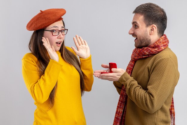 young beautiful couple happy man making proposal with engagement ring in red box to his confused girlfriend in beret during valentines day standing over white background