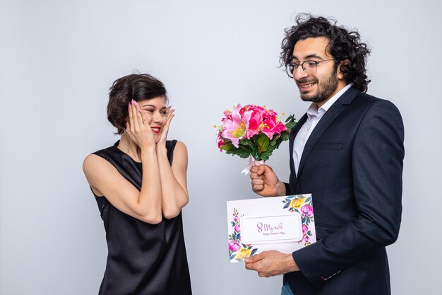 Young beautiful couple happy man with greeting card giving a bouquet of flowers to his surprised and happy girlfriend celebrating international women's day march 8