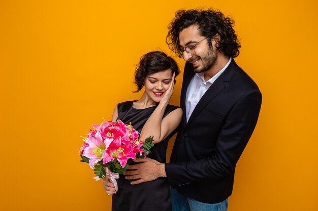 Young beautiful couple happy man and woman with bouquet of flowers smiling cheerfully embracing happy in love celebrating international women's day march 8 standing over orange background