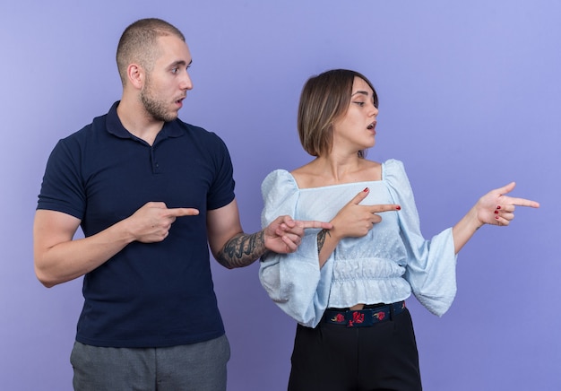 Free photo young beautiful couple man and woman looking aside intrigued pointing with index fingers to the side standing over blue wall