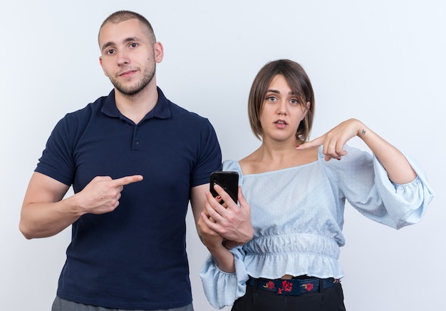 Young beautiful couple man and woman looking man with smartphone pointing with index finger at his confused girlfriend standing