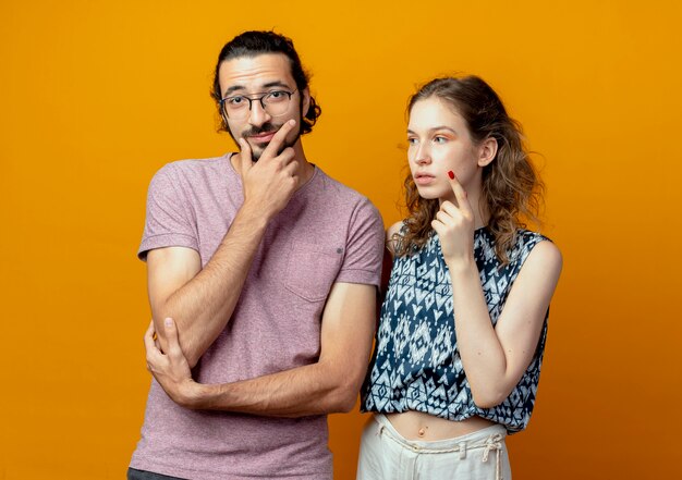 Young beautiful couple man and women looking puzzled thinking standing over orange background