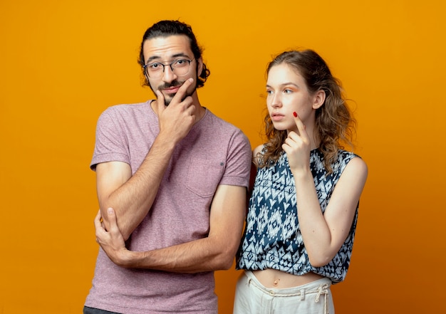 young beautiful couple man and women looking puzzled thinking standing over orange wall