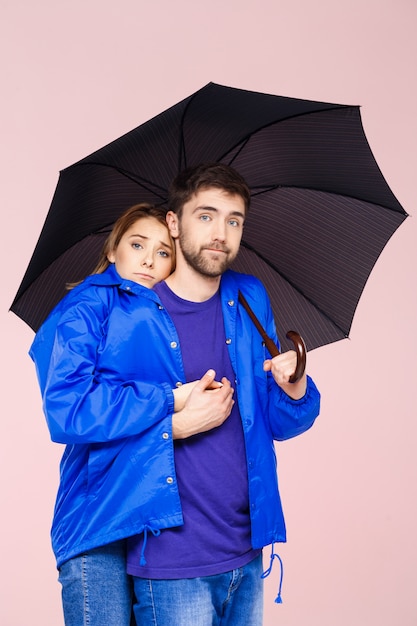 Young beautiful couple posing wearing one rain coat holding umbrella over light pink wall