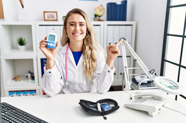 Young beautiful doctor woman holding glucose meter smiling happy pointing with hand and finger to the side