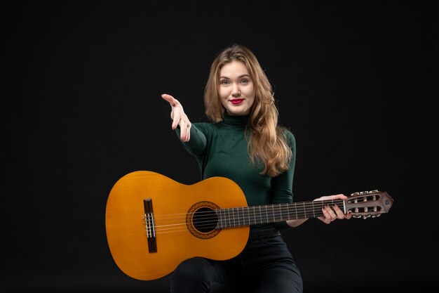 Young beautiful female guitarist holding her favorite musical instrument elcoming someone on dark