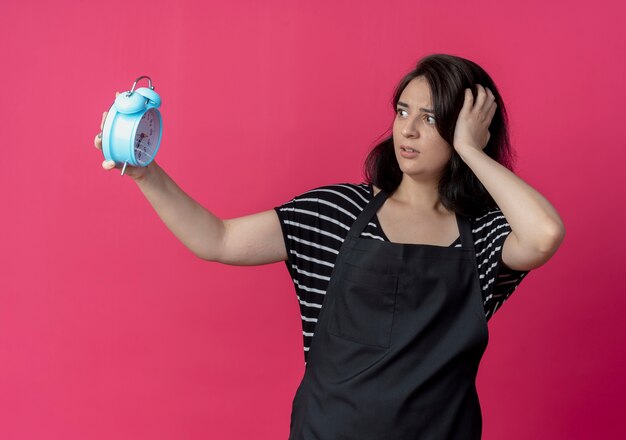 Young beautiful female hairdresser in apron holding alarm clock looking at it confused and worried standing over pink wall