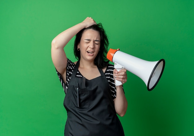 Young beautiful female hairdresser in apron holding megaphone looking confused with hand on her head standing over green wall