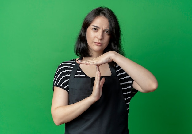 Young beautiful female hairdresser in apron lookign at camera with serious face showing time out gesture  over green