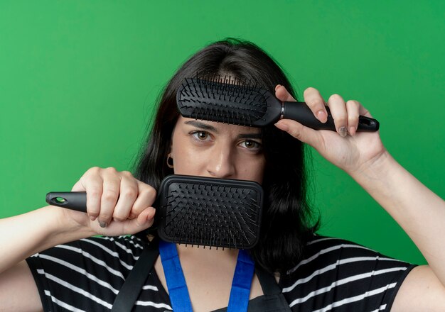 Young beautiful female hairdresser in apron with gold medal around neck holding two hair brushes in front of her face  with serious expression standing over green wall