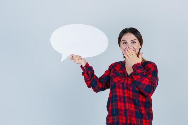 Young beautiful female holding speech bubble while covering mouth with hand in casual shirt, jeans and looking blissful. front view.