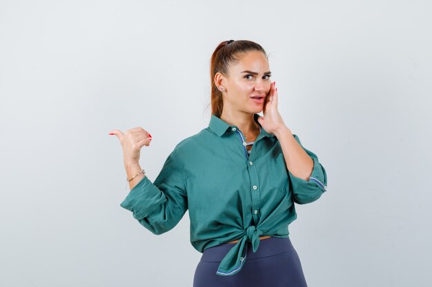 Young beautiful female pointing left with thumb while keeping hand near mouth in green shirt and looking curious , front view.