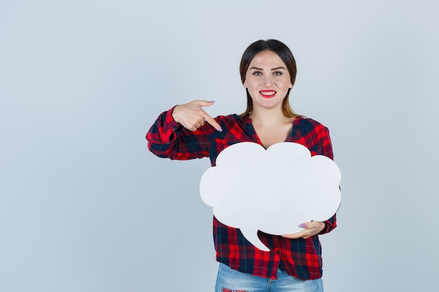 Young beautiful female pointing at speech bubble in casual shirt, jeans and looking cheerful. front view.