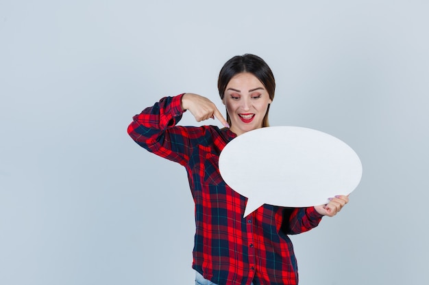 Young beautiful female pointing at speech bubble in casual shirt, jeans and looking merry. front view.