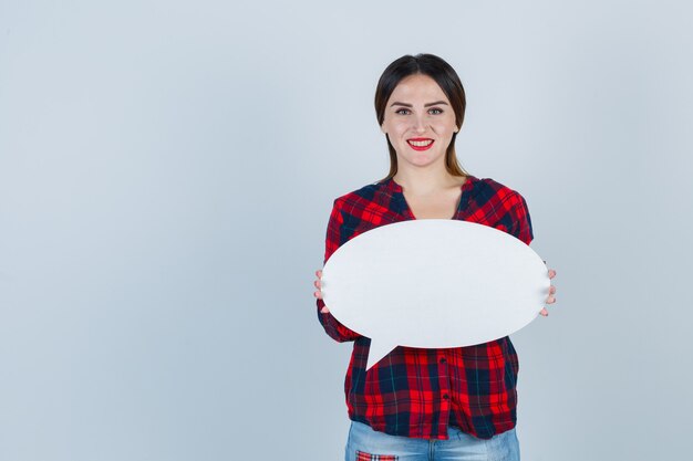 Young beautiful female posing while holding speech bubble in casual shirt, jeans and looking positive , front view.