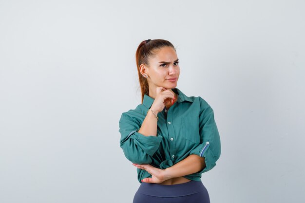 Young beautiful female standing in thinking pose in green shirt and looking puzzled. front view.