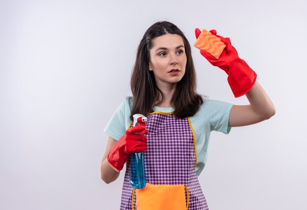 Young beautiful girl in apron and rubber gloves holding cleaning spray and sponge going to clean looking confident 