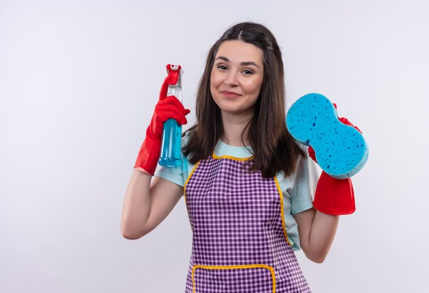 Young beautiful girl in apron and rubber gloves holding cleaning spray and sponge looking at camera with happy face smiling 